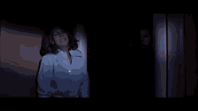a woman is standing in a dark room in front of a door with a ghost behind her .