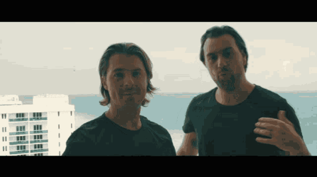 two men standing next to each other in front of a building with the ocean in the background