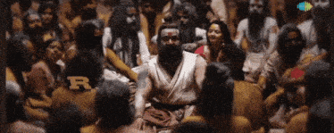 a man with a beard sits in the middle of a crowd of people with the letter r visible