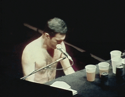 a shirtless man singing into a microphone while sitting at a table with plastic cups
