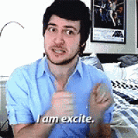 a man in a blue shirt is standing in front of a bed and says `` i am excite '' .