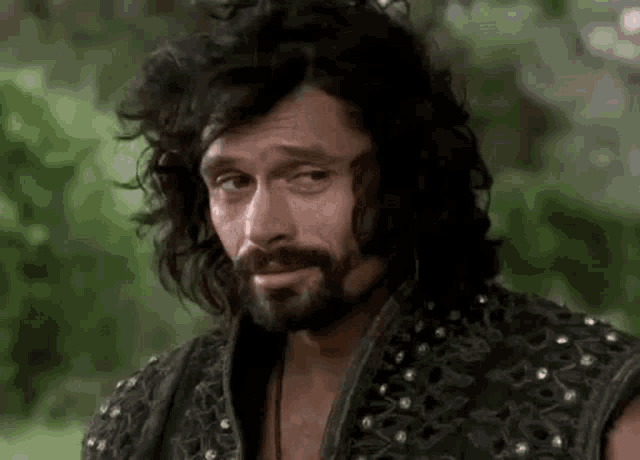 a man with long curly hair and a beard is wearing a black vest .