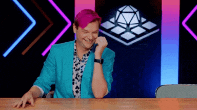 a man with pink hair and a blue jacket is sitting at a table with his hands on his face .