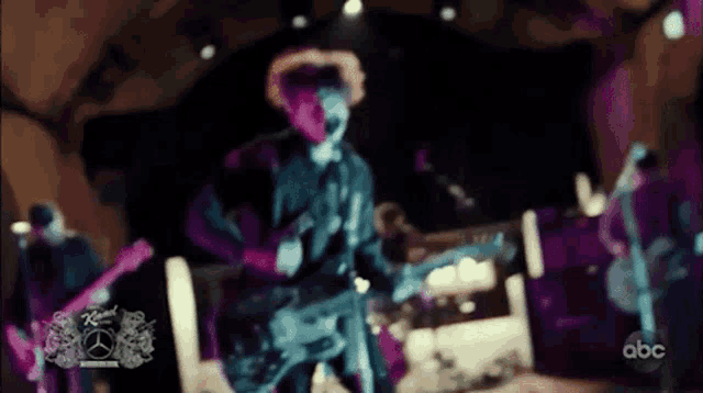 a blurry picture of a man playing a guitar with a mercedes logo on the bottom right