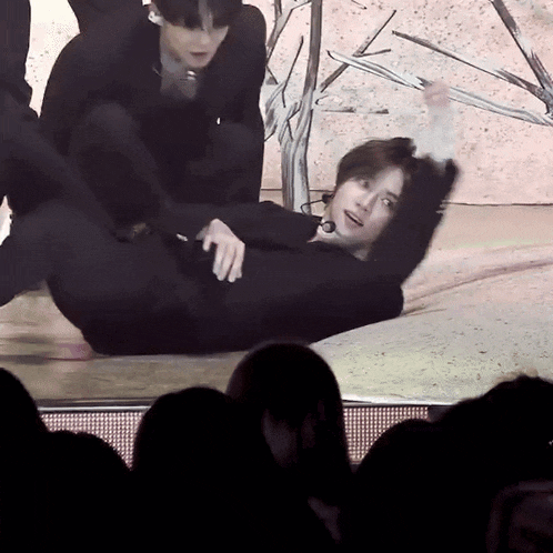 a man in a black suit is laying on his back on a stage while another man stands behind him