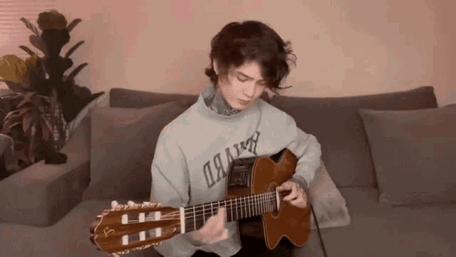 a young man is playing an acoustic guitar on a couch while wearing a sweatshirt that says oakland
