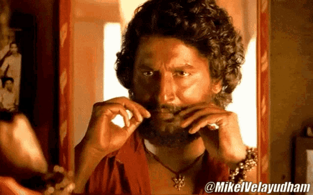 a man with a beard is looking at himself in a mirror with @mikelvelayudham written on the bottom