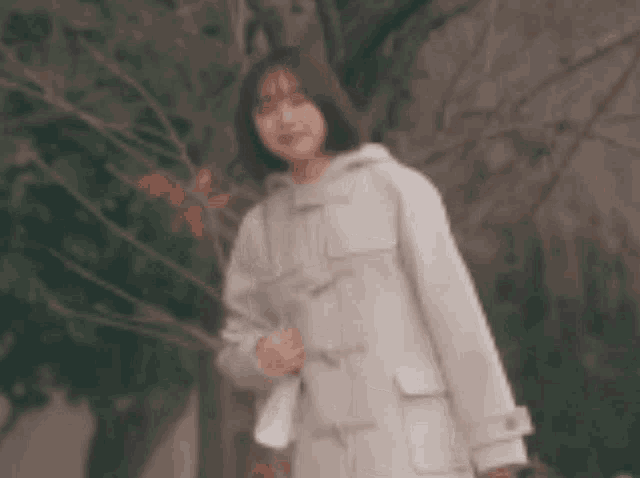 a woman in a white coat is standing in front of a tree and waving .