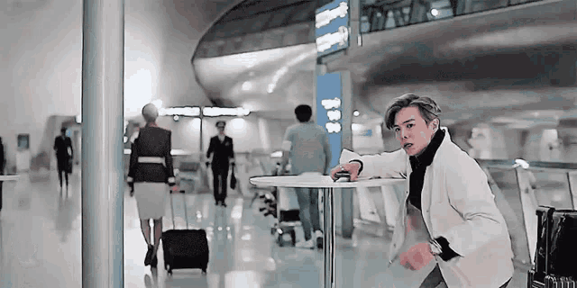 a man is leaning on a table in an airport while a woman walks with a suitcase .