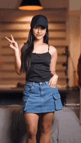 a woman wearing a black tank top and a denim skirt is standing in a room giving a peace sign .