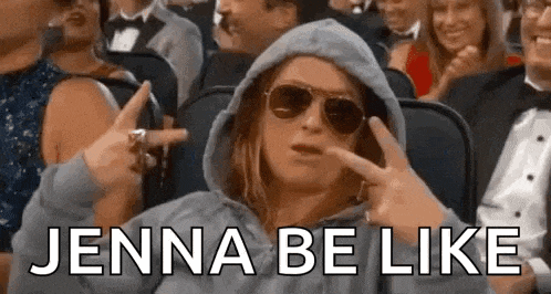 a woman wearing a hoodie and sunglasses is sitting in a crowd of people and making a peace sign .