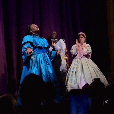 a woman in a blue dress stands on a stage in front of a purple curtain