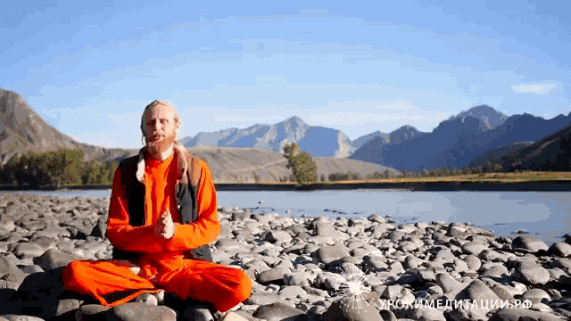 a man sits on rocks near a river with mountains in the background and the words " урокмедитации.рф " below him
