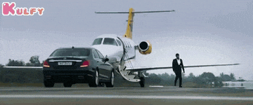 a man in a tuxedo walks next to a car and an airplane on a runway .