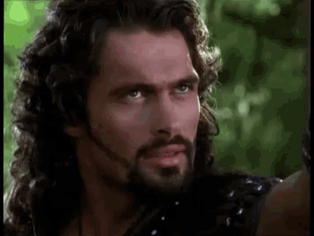 a man with long hair and a beard is standing in the woods and looking at the camera .