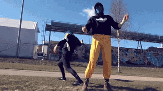 a man wearing a black adidas sweatshirt stands next to another man wearing yellow pants