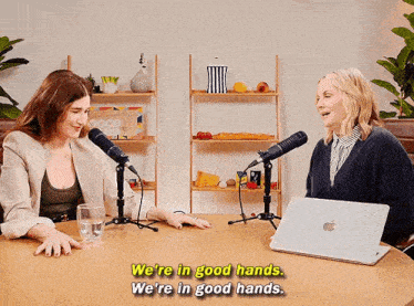 two women are sitting at a table with microphones and a laptop and one of them is saying we 're in good hands .