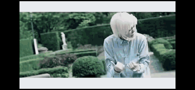 a man with white hair is standing in a garden with a statue in the background .