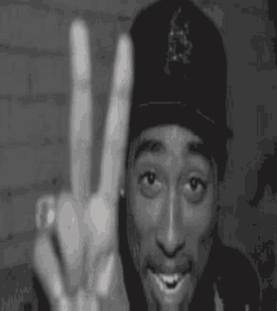 a black and white photo of tupac shakur making a peace sign with his hands .