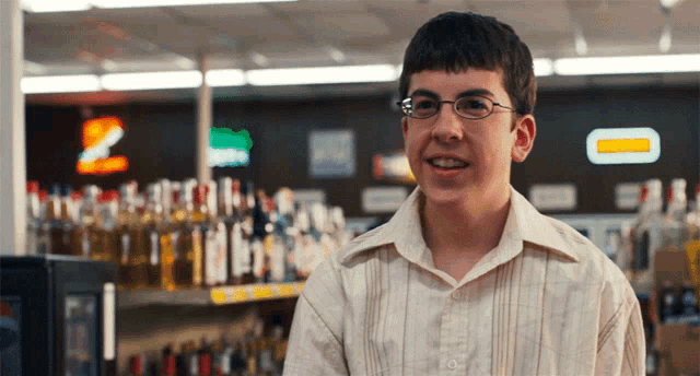 a young man wearing glasses is smiling in front of a shelf of liquor