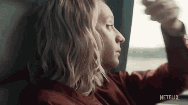 a woman in a red shirt is looking out a window with netflix written on the bottom right