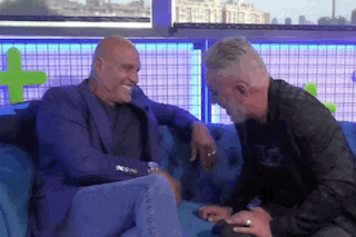 two men are sitting on a blue couch talking to each other and smiling .