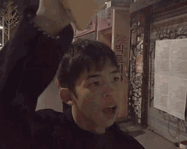 a man stands in front of a pink building with chinese writing on it and says hi