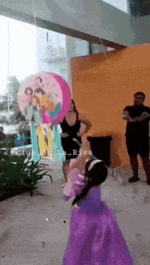 a little girl in a purple dress is holding a piñata