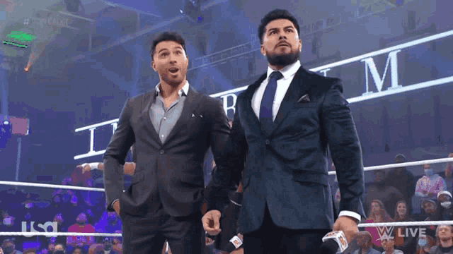 two men in suits are standing in front of a wrestling ring that says tbm on it