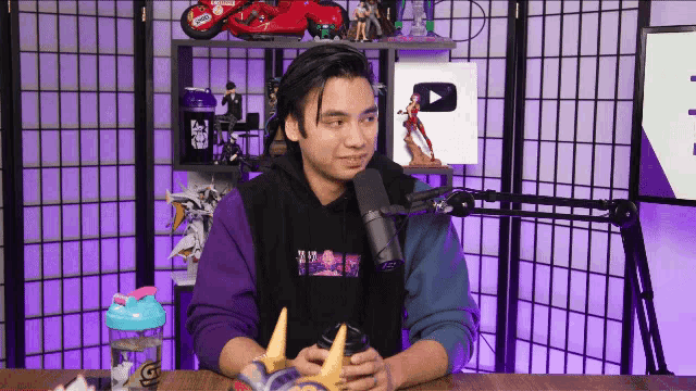 a man in a purple hoodie is sitting in front of a microphone in a room .