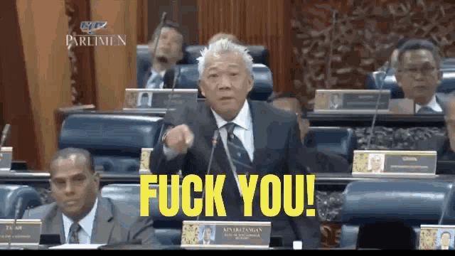 a man in a suit and tie is giving a speech in front of a group of people and the words fuck you are on the screen