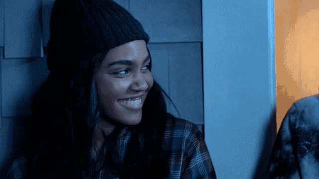a woman wearing a black beanie and plaid shirt smiles in a dark room