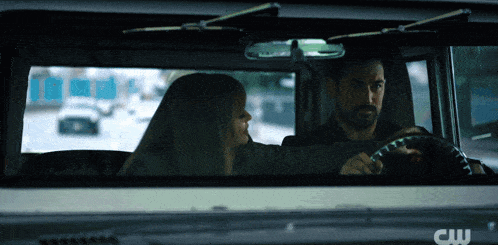 a man and a woman are sitting in a car with the cw logo on the hood