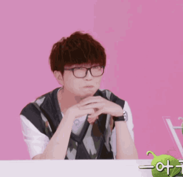 a man wearing glasses sits in front of a pink background with a green sticker that says " a "