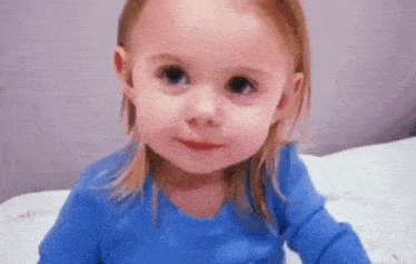 a little girl is sitting on a bed wearing a blue shirt and looking at the camera .