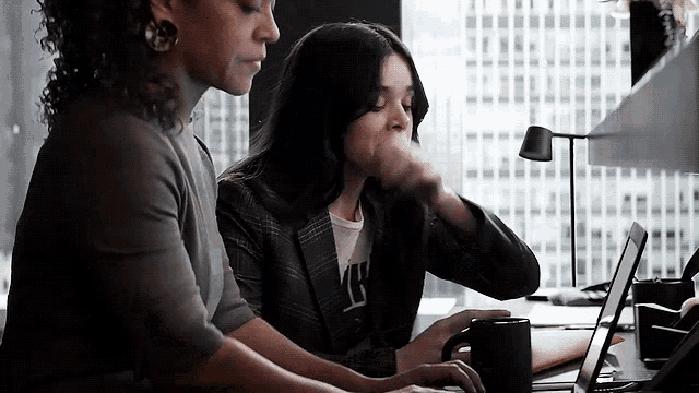 two women are sitting at a desk with a laptop computer .