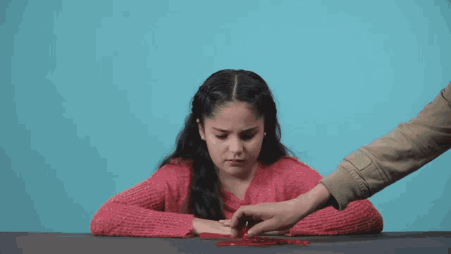 a girl in a pink sweater is sitting at a table with a hand reaching out towards her