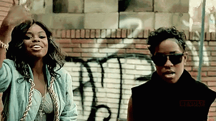 two women standing next to each other in front of a brick wall with graffiti on it .