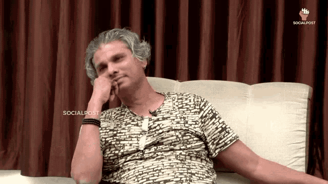 a man is sitting on a couch with his hand on his chin and a socialpost logo behind him