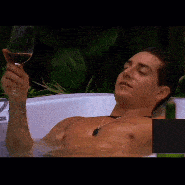 a shirtless man is laying in a bathtub holding a glass of red wine