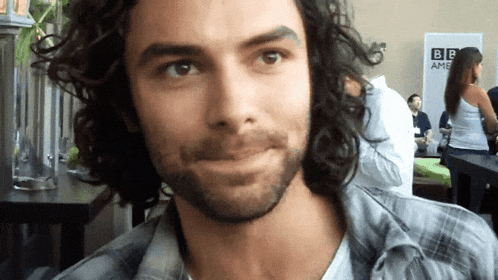 a man with curly hair and a beard looks at the camera in front of a bbc sign