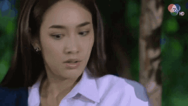 a woman in a white shirt is standing in front of a tree and looking at the camera .
