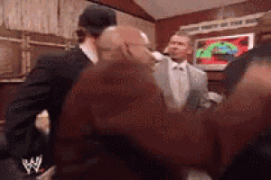 a group of men are standing in a room and one of them is wearing a wrestling logo on his back .