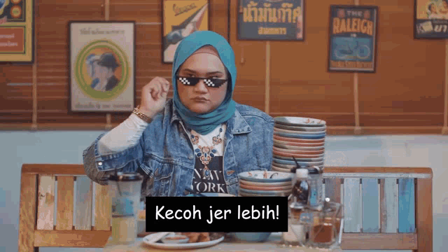 a woman wearing a hijab and sunglasses is sitting at a table with plates of food and says kecoh jer lebih
