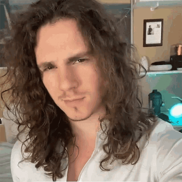 a man with long curly hair is wearing a white shirt and looking at the camera
