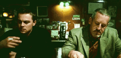 two men are sitting at a table in a bar smoking a cigarette and drinking beer .