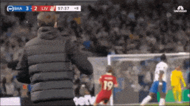 a man in a black vest is standing on a soccer field watching a soccer game being played .