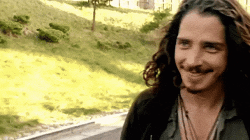 a man with long hair and a beard is smiling in front of a field of grass .