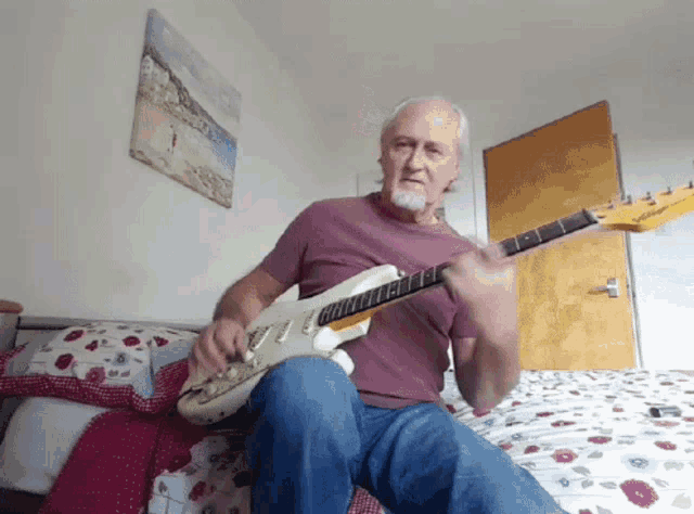 a man playing a guitar on a bed with a picture of a beach on the wall behind him