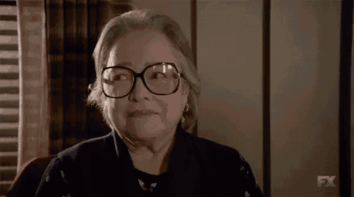 an elderly woman wearing glasses is sitting in front of a window and says next !
